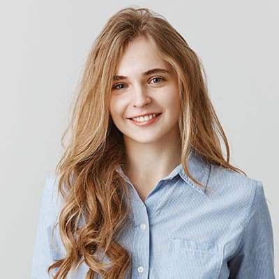 A person with long, wavy hair and a smiling expression is wearing a light blue button-up shirt. The background is plain and neutral.