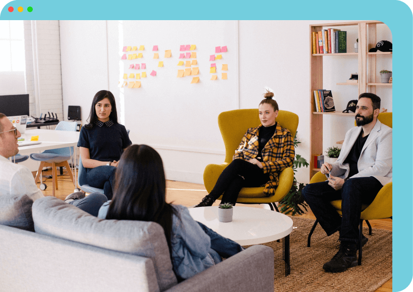 A group of people sitting in an office space, engaged in discussion. Two individuals are seated on a couch, while three others sit on armchairs. The background features a whiteboard with sticky notes and a bookshelf.