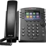 A black Polycom VoIP office telephone features a digital display with colorful icons. It includes a corded handset, number keypad, and multiple buttons for navigation and functions. The phone is angled to the right, offering a sleek blend of technology and style.