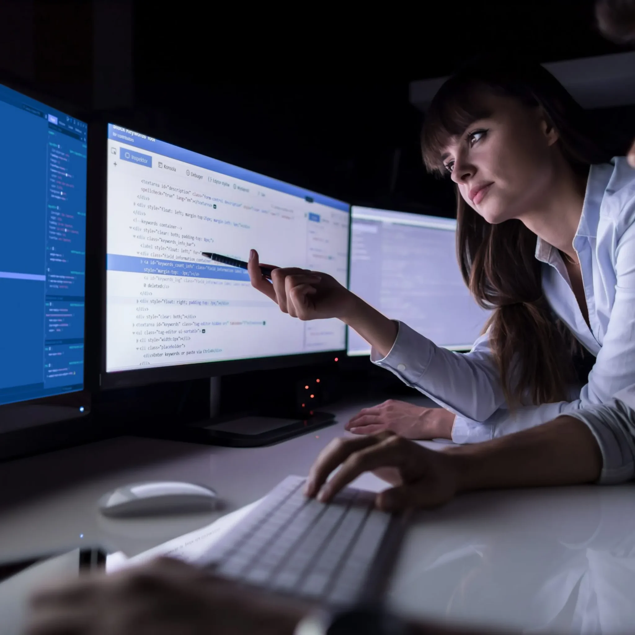 In a dimly lit room filled with multiple monitors, a woman in a white shirt points at a computer screen displaying code, diving into website development. Her collaboration with a colleague typing on the keyboard is crucial for driving business success.